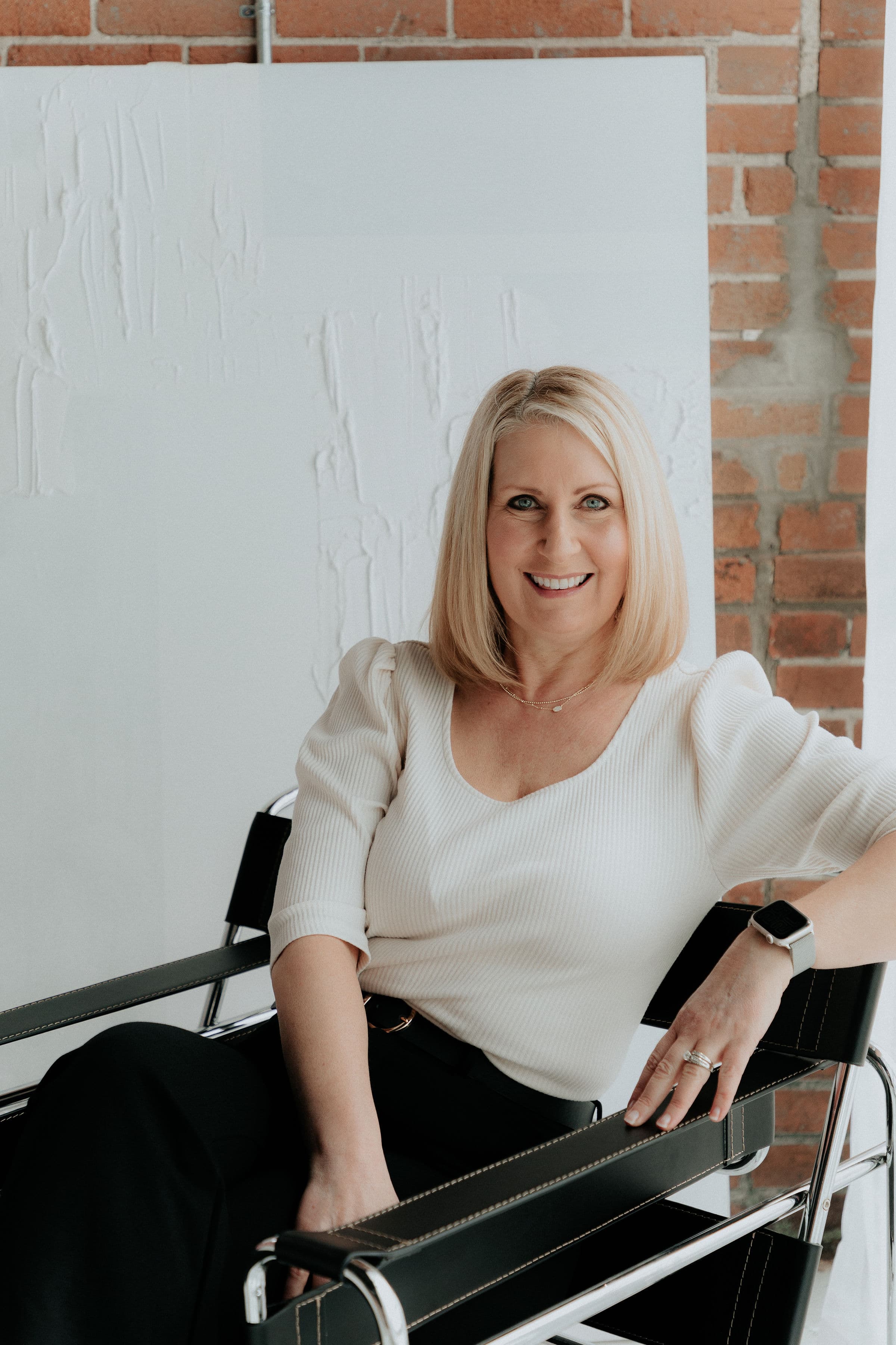 Smiling blonde woman in a white puff-sleeve top sitting in a modern black leather chair.